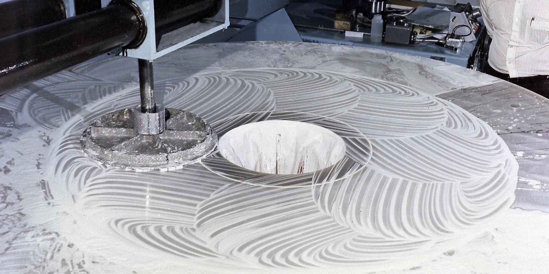 A large telescope mirror under a polishing tool, with circular polishing traces across its surface.